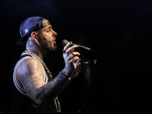 Cantor tatuado com boné e microfone em palco escuro.