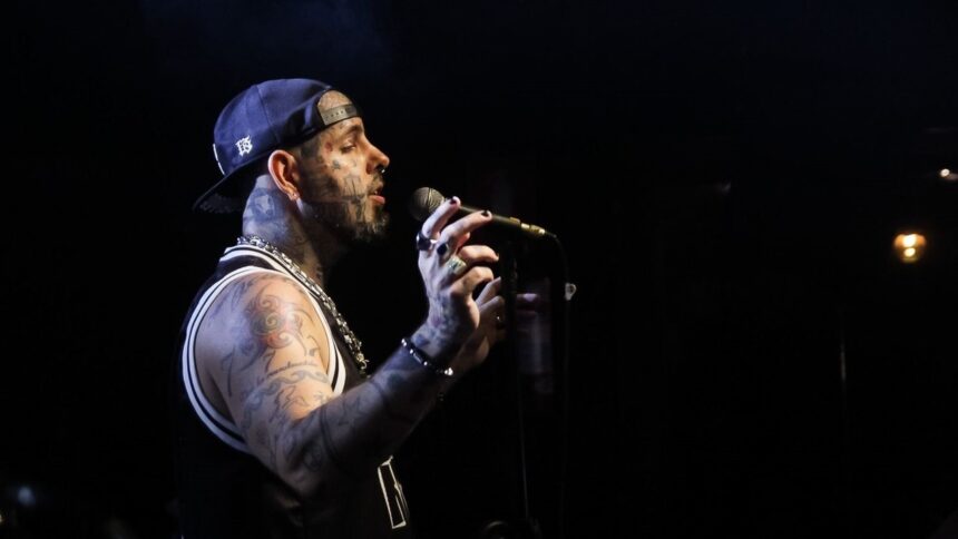 Cantor tatuado com boné e microfone em palco escuro.