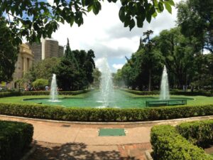 Fontes em parque urbano com gramados, árvores e prédios ao fundo sob céu nublado.