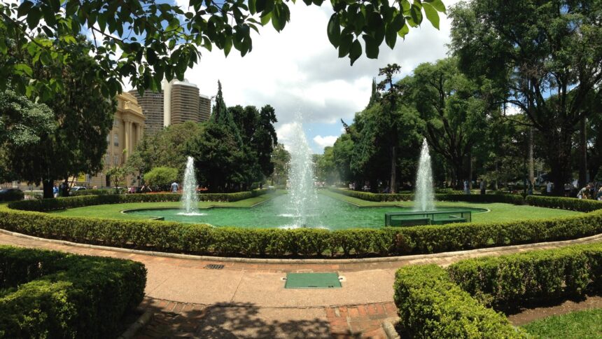 Fontes em parque urbano com gramados, árvores e prédios ao fundo sob céu nublado.