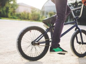 Pernas de pessoa em bicicleta azul escura, tênis verde, pedais, em área externa pavimentada.