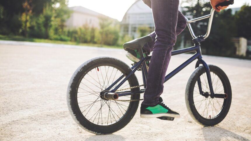 Pernas de pessoa em bicicleta azul escura, tênis verde, pedais, em área externa pavimentada.