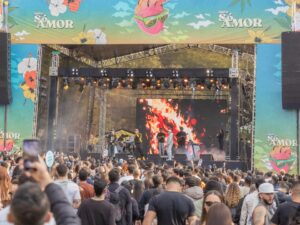 Músicos se apresentam em palco de festival com telão de chamas e público filmando, banners 'Só Amor'.