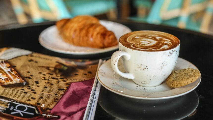 Xícara branca de café com latte art e biscoito, ao lado de um croissant e revista em mesa.