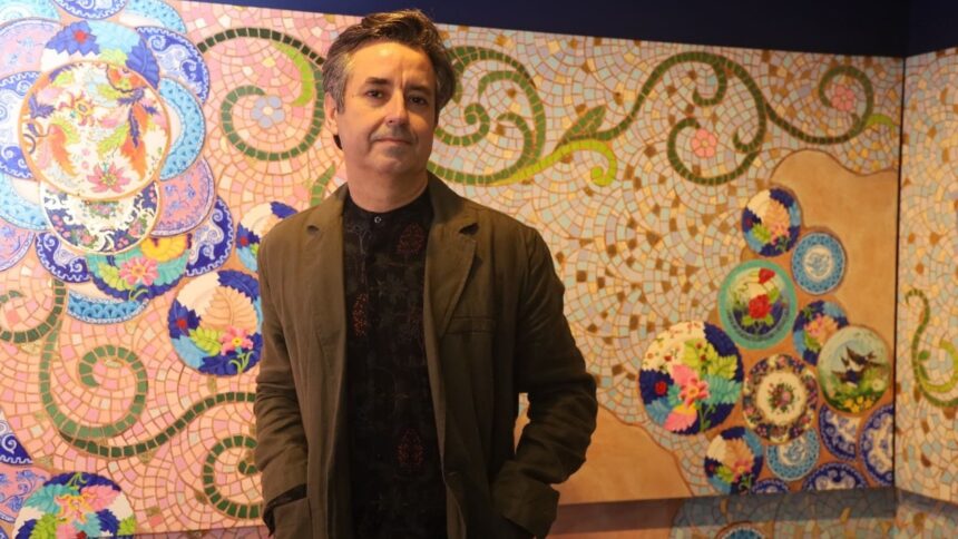 Iuri Sarmento em pé diante de painel de mosaico colorido com elementos florais.
