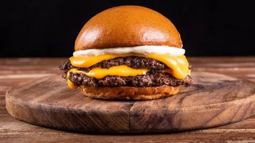 Hambúrguer duplo com pão brioche, dois queijos derretidos e molho branco em tábua de madeira.