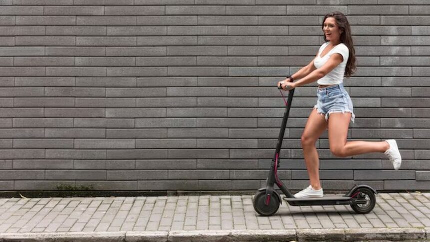 Jovem mulher sorridente impulsiona patinete elétrico em calçada de paralelepípedos com parede cinza ao fundo.