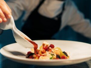 Mão de chef regando sobremesa colorida com calda vermelha em prato branco.