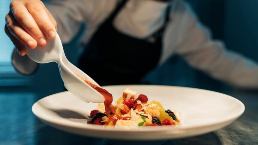 Mão de chef regando sobremesa colorida com calda vermelha em prato branco.