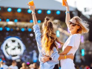 Duas mulheres em festival, braços erguidos com copos, palco iluminado no fundo.