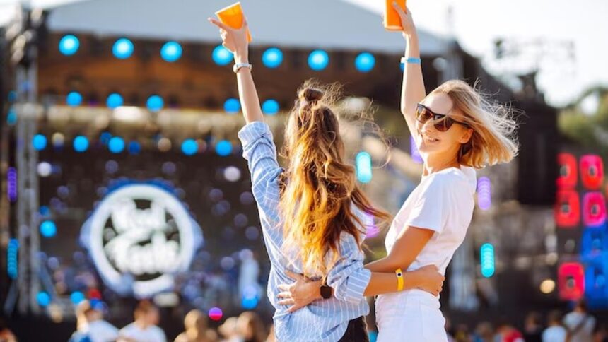 Duas mulheres em festival, braços erguidos com copos, palco iluminado no fundo.