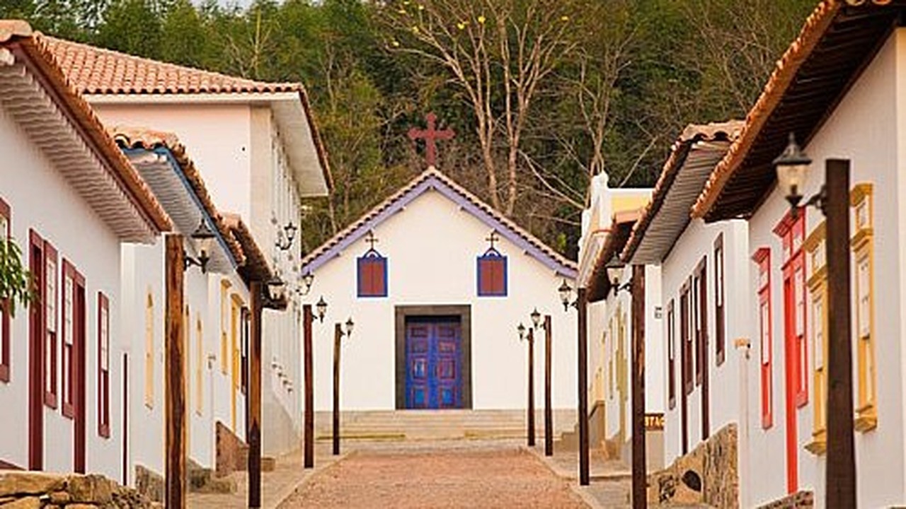 Pequena igreja branca com porta azul e cruz vermelha, ladeada por casas coloniais.