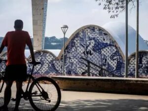 Ciclista de costas com bicicleta em frente à Lagoa da Pampulha e arquitetura azul e branca.
