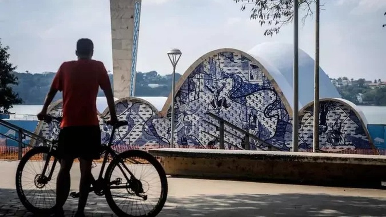 Ciclista de costas com bicicleta em frente à Lagoa da Pampulha e arquitetura azul e branca.