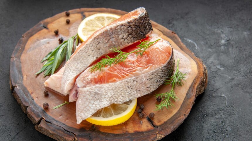 Dois filés de salmão cru com pele, fatias de limão e ervas frescas em tábua de madeira.