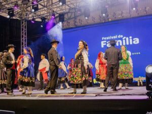 Grupo de pessoas com trajes folclóricos coloridos em palco iluminado com tela azul ao fundo.