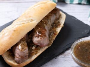 Choripán com linguiça e molho chimichurri em pão francês, servido em tábua preta, com tigela de molho ao lado.