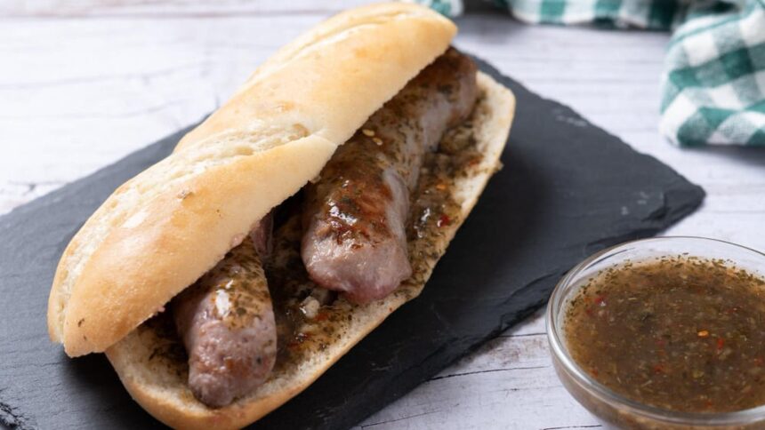 Choripán com linguiça e molho chimichurri em pão francês, servido em tábua preta, com tigela de molho ao lado.