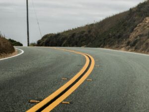 Estrada de asfalto curvada com faixa dupla amarela, tachas refletoras, encostas e céu nublado.
