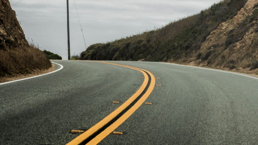 Estrada de asfalto curvada com faixa dupla amarela, tachas refletoras, encostas e céu nublado.