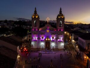 Igreja histórica com iluminação roxa e dourada, cruz brilhante, e pessoas em praça noturna.
