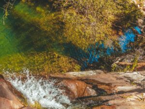 Vista aérea de água cristalina verde-azulada e queda d'água entre rochas e vegetação.