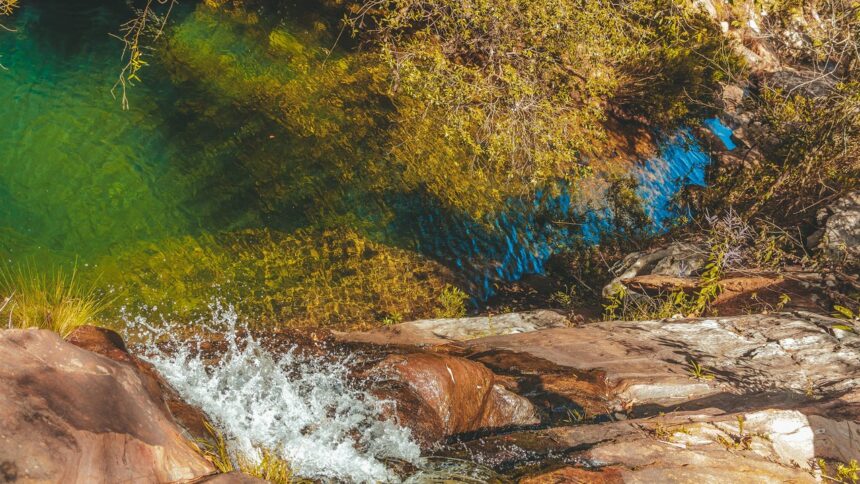 Vista aérea de água cristalina verde-azulada e queda d'água entre rochas e vegetação.