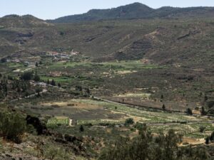 Vista aérea de vale montanhoso com campos cultivados, vilarejo e estradas sinuosas sob céu claro.