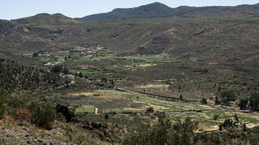 Vista aérea de vale montanhoso com campos cultivados, vilarejo e estradas sinuosas sob céu claro.