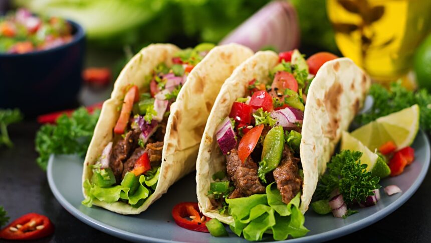 Três tacos recheados com carne, alface, tomate, cebola roxa e pimentão, servidos com limão.