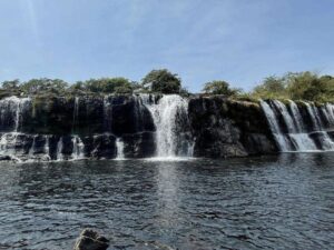 Múltiplas quedas d'água de uma cachoeira rochosa com vegetação descem para um rio escuro.