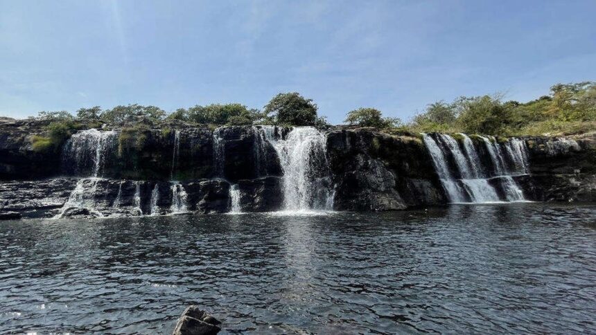 Múltiplas quedas d'água de uma cachoeira rochosa com vegetação descem para um rio escuro.