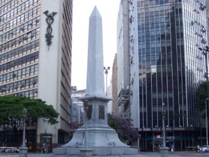 Obelisco branco em praça urbana cercado por prédios altos, pessoas e árvores.