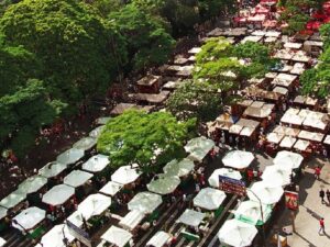 Vista aérea de um mercado ao ar livre com várias barracas brancas, árvores e pessoas.