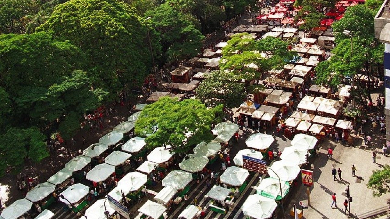 Vista aérea de um mercado ao ar livre com várias barracas brancas, árvores e pessoas.