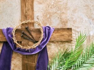 Cruz de madeira com pano roxo, coroa de espinhos, pregos e ramos de palmeira.
