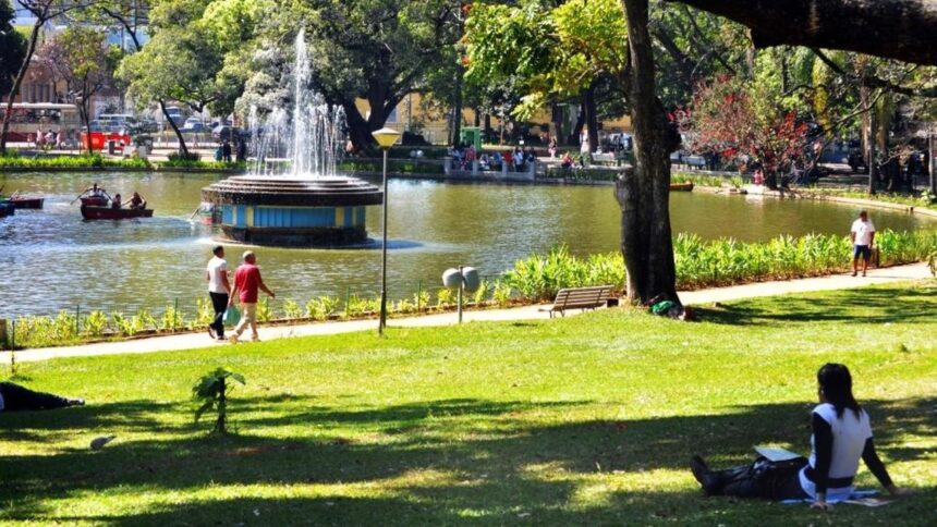 Lago com fonte, pessoas em barcos e caminhando, gramado e árvores em parque.