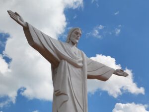 Estátua branca de Jesus Cristo de braços abertos contra um céu azul com nuvens brancas.