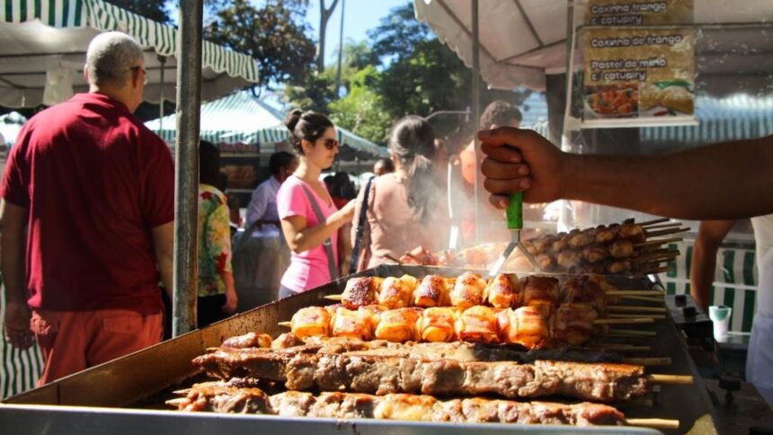 Mão grelhando espetinhos de carne e bacon em feira com pessoas e barracas de comida.