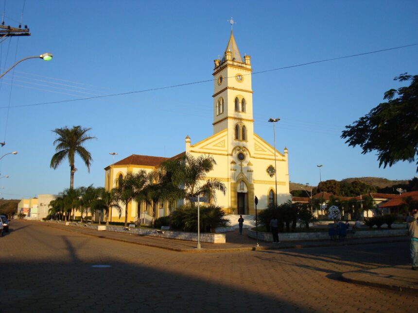 Igreja amarela com torre, relógio, cruzeiro, palmeiras, rua e céu azul claro.