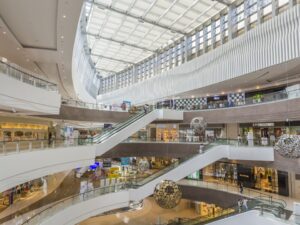 Shoppings center multiandares com várias escadas rolantes, lojas e pessoas circulando sob teto de vidro.