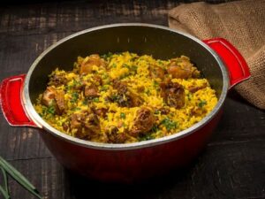 Panela vermelha com galinhada, arroz amarelo, pedaços de frango, cheiro-verde, em mesa de madeira rústica.
