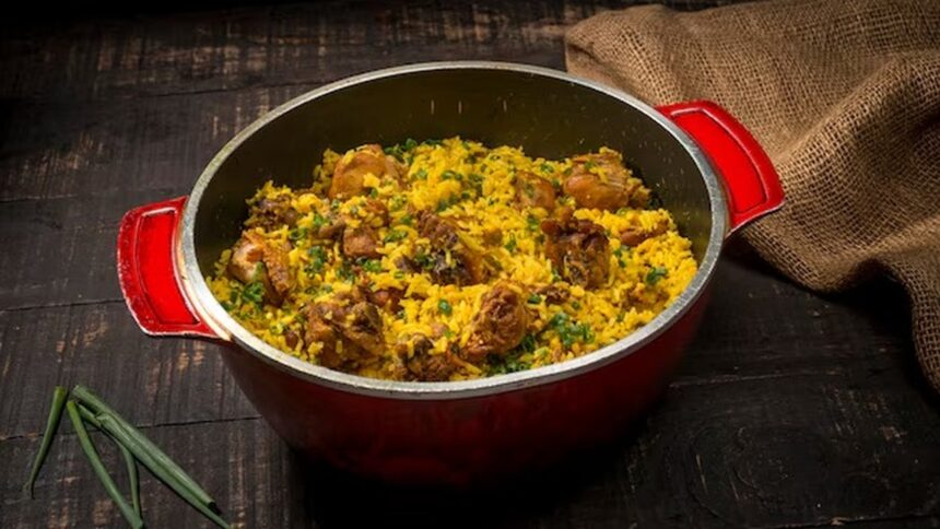 Panela vermelha com galinhada, arroz amarelo, pedaços de frango, cheiro-verde, em mesa de madeira rústica.