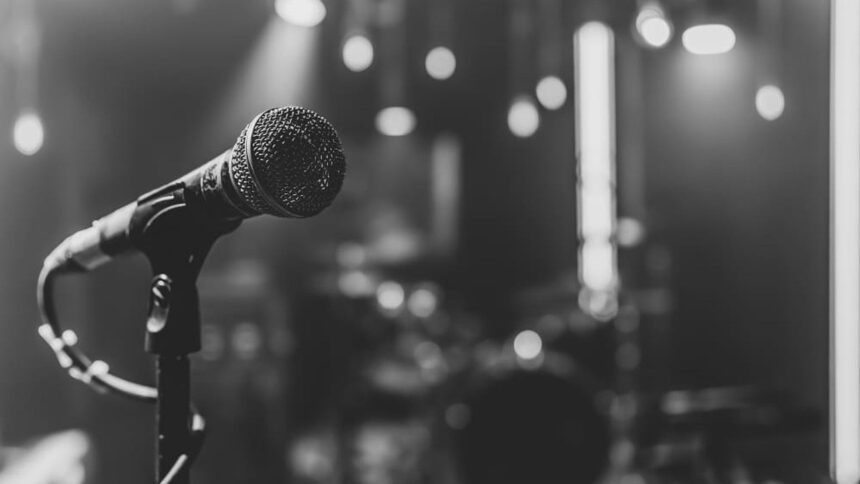 Microfone preto em foco no palco escuro com luzes desfocadas, em preto e branco.