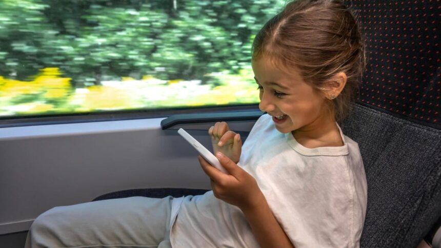 Menina sorridente usando smartphone branco em assento de trem, com paisagem borrada ao fundo.