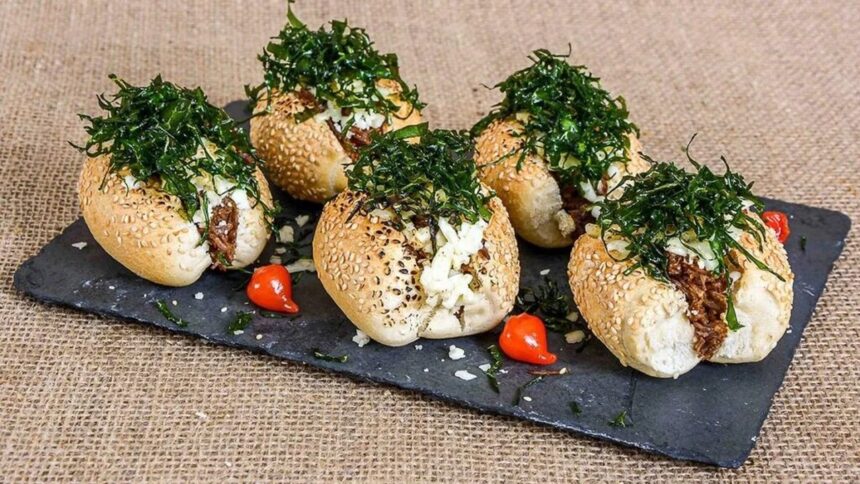Cinco pães com gergelim recheados com carne, queijo e couve, servidos em ardósia com pimentas.