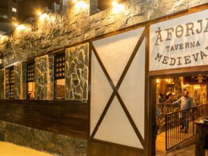 Fachada noturna A Forja Taverna Medieval iluminada, com letreiro, entrada e pessoas dentro.