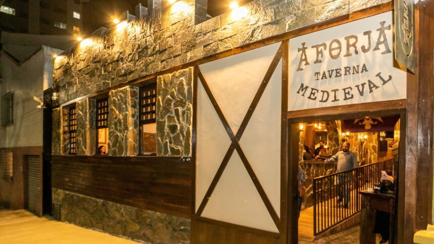 Fachada noturna A Forja Taverna Medieval iluminada, com letreiro, entrada e pessoas dentro.