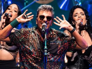 Homem de óculos escuros e duas mulheres cantam em microfones no palco, sob luzes azuis.