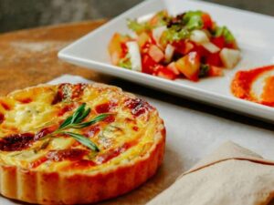 Quiche individual dourada com ramo de alecrim, acompanhada de salada e molho em um prato branco.